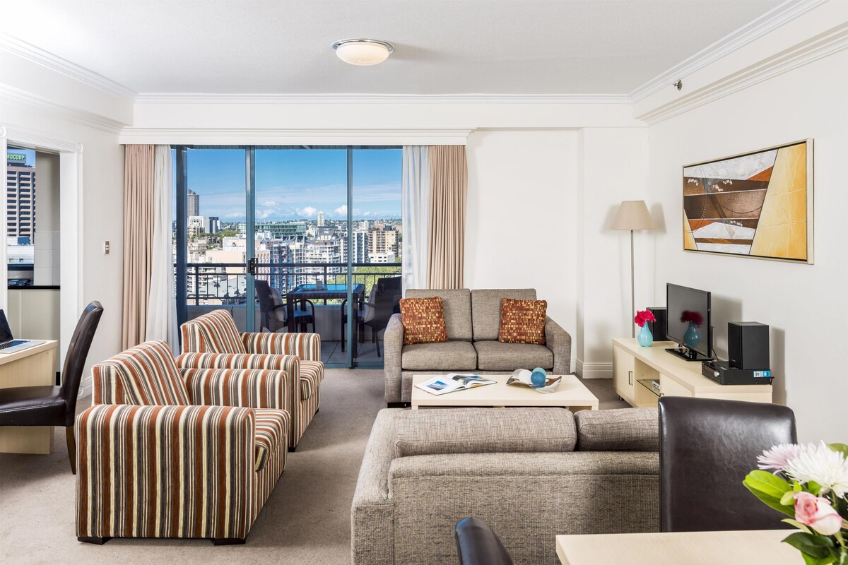 Habitación de hotel con vista a la ciudad, un sofá, una mesa de comedor, un televisor y una lámpara.