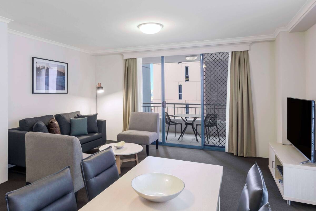 A modern hotel room with a sofa, chairs, a dining table, a TV, and a balcony with a view.