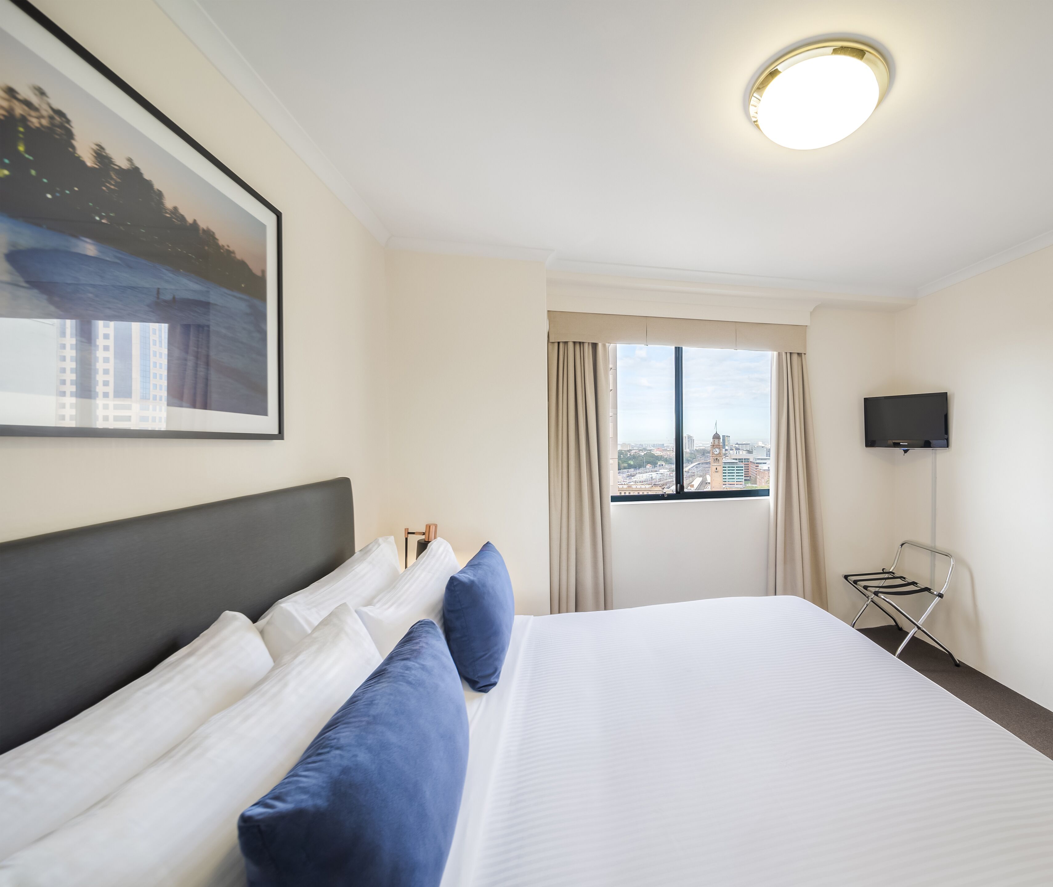 A hotel room with a large bed, a flat-screen TV, a window with a city view, and a ceiling light.
