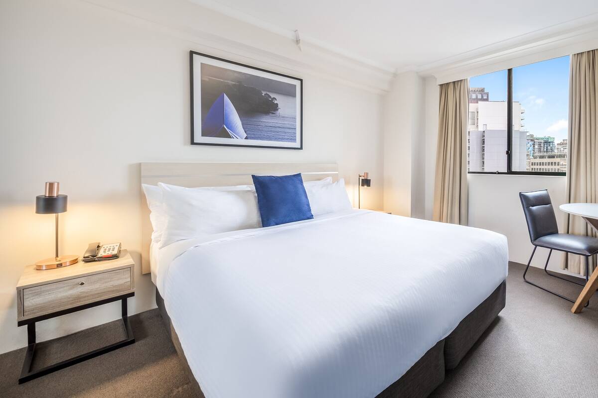 A hotel room with a large bed, a bedside table with a lamp, a chair, and a view of the city through a window.