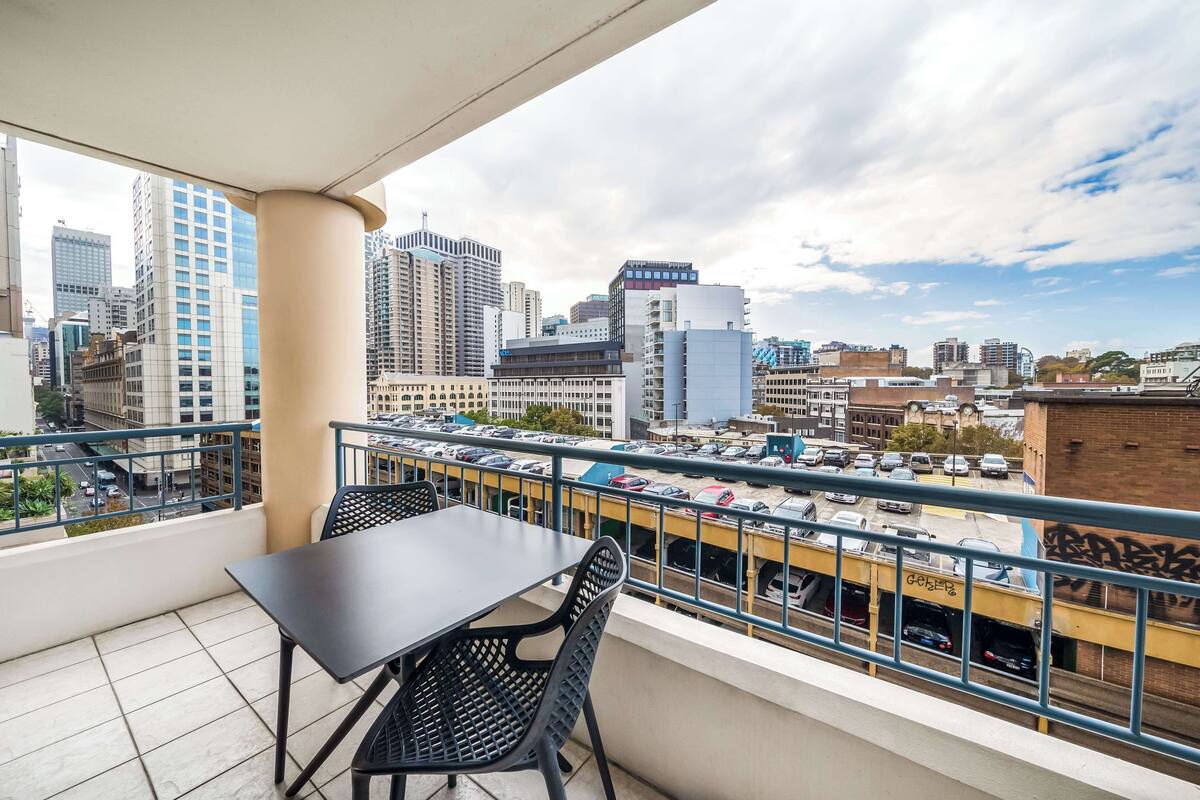 A balcony with a table and chairs, overlooking a cityscape with tall buildings and a parking area.