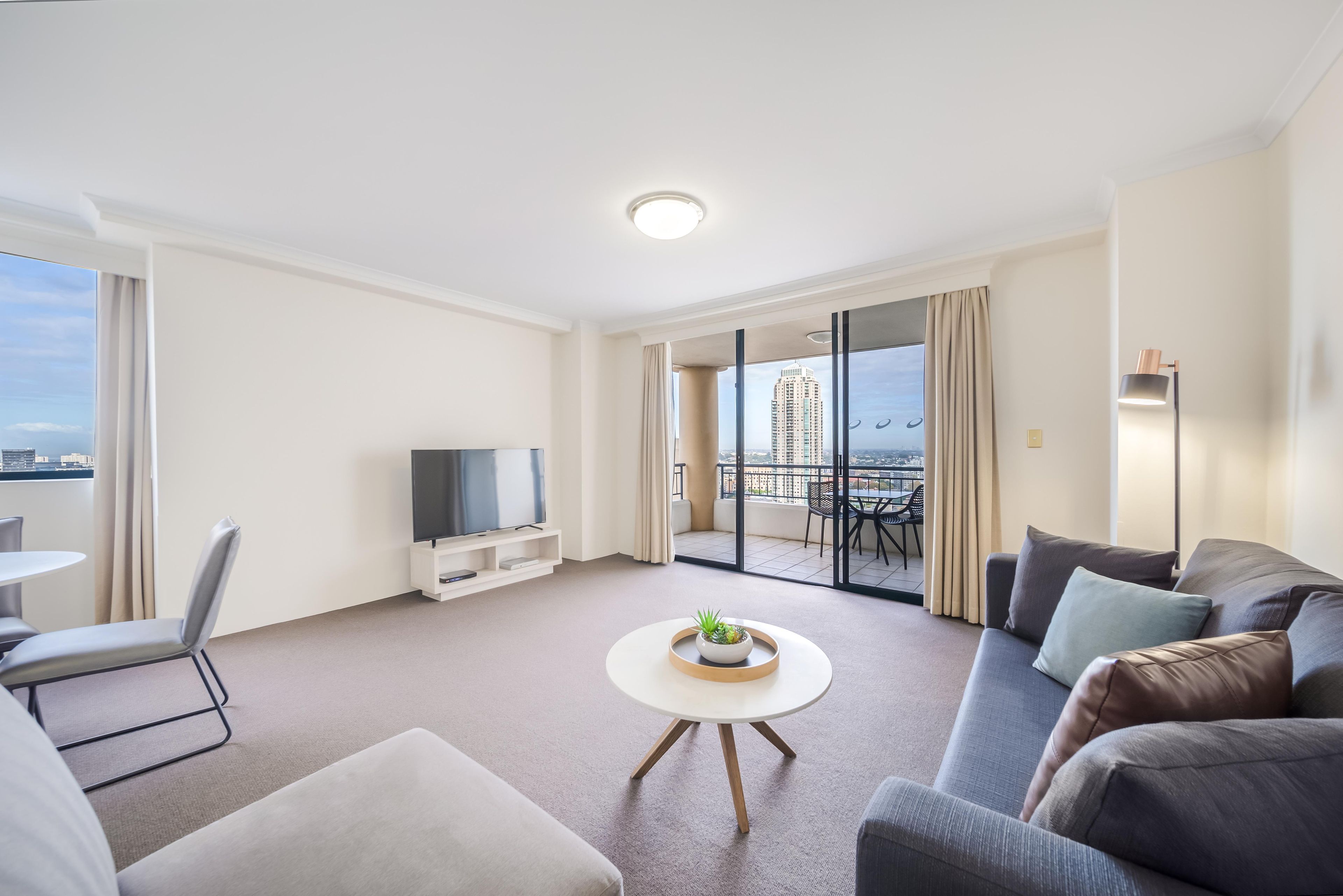 Een moderne woonkamer met een flatscreen televisie, een ronde salontafel met een plant, een bank met kussens, een eethoek met een tafel en stoelen, en een balkon met uitzicht op de stad.