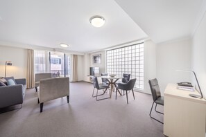 Habitación de hotel con sofá, mesa de comedor con sillas, escritorio con lámpara y ventanas amplias.