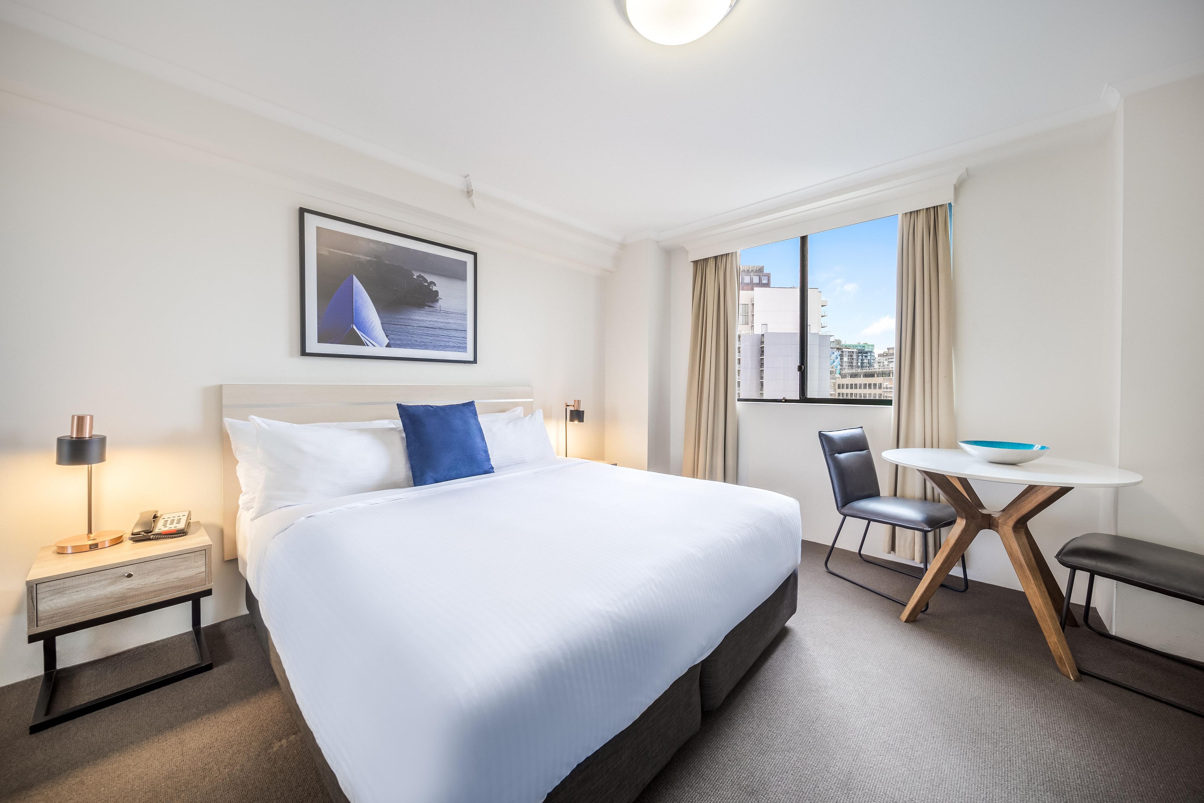 Een hotelkamer met een groot bed, een bureau met een stoel, een kleine tafel met een kom, een nachtkastje met een lamp en een lijst aan de muur.