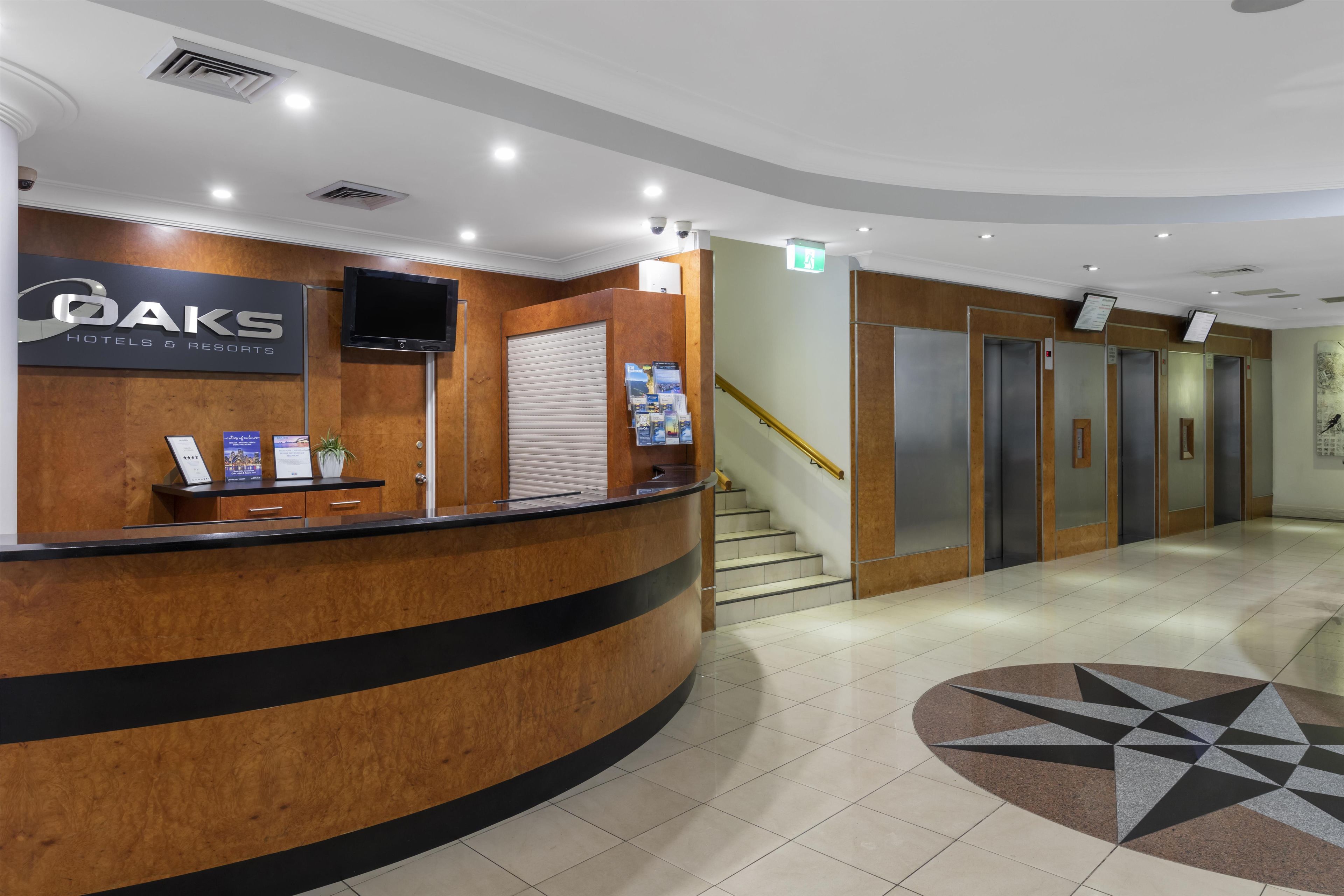 A hotel reception desk with a sign for OAKS Hotels & Resorts, a staircase, and a set of elevators.