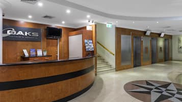 A hotel reception desk with a sign for OAKS Hotels & Resorts, a staircase, and a set of elevators.