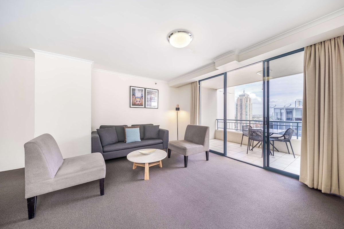 A hotel room with a grey sofa, a grey armchair, a small round table, and a balcony with outdoor seating.