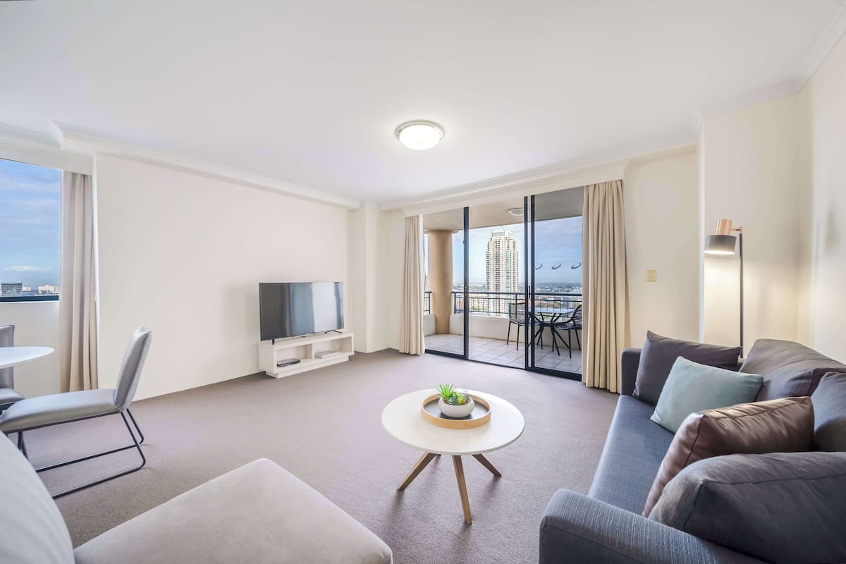 A modern living room with a sofa, a round coffee table with a plant, a TV, and a balcony with a city view.