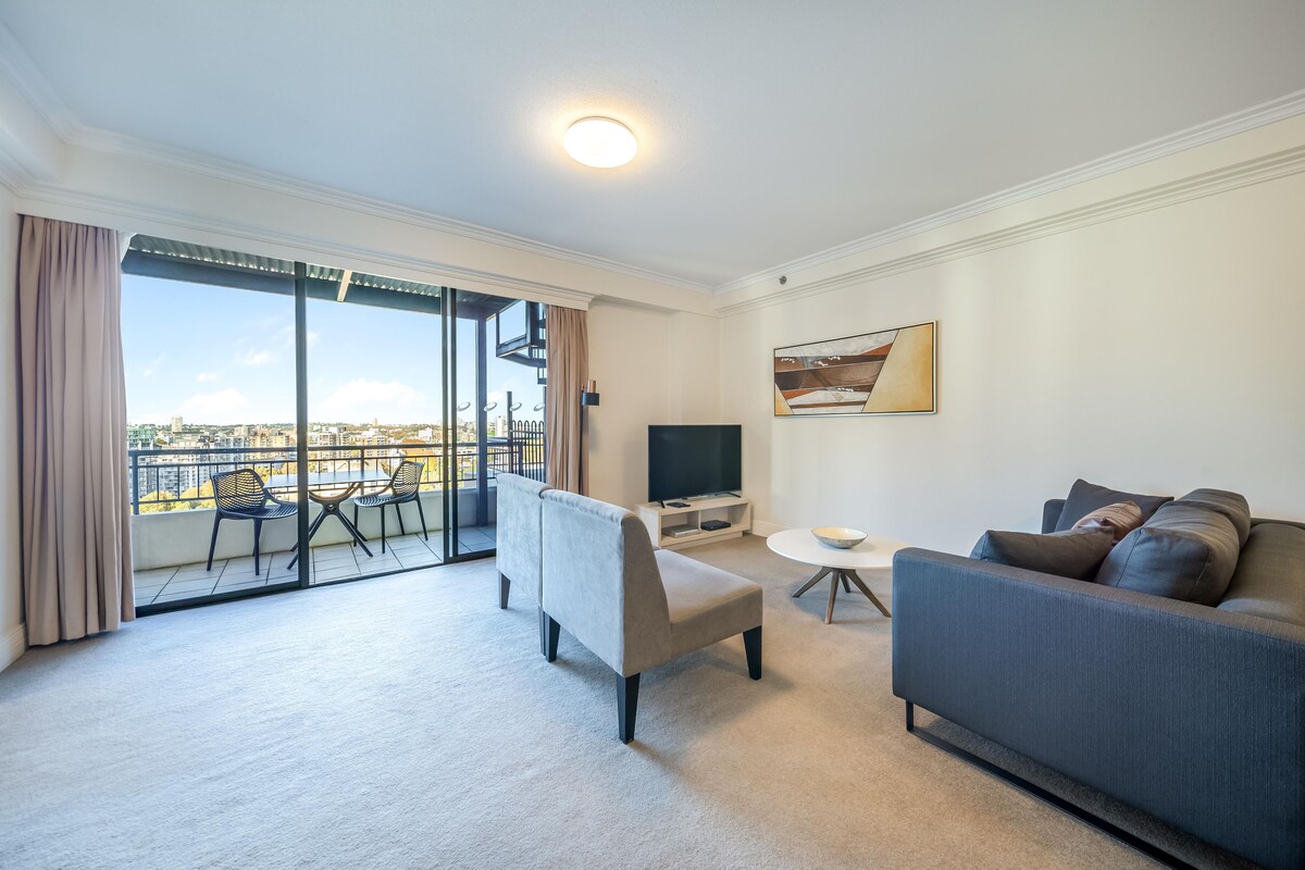 Una sala de estar moderna con un sofá, un sillón y una mesa de comedor con sillas. Hay un televisor y un cuadro en la pared.
