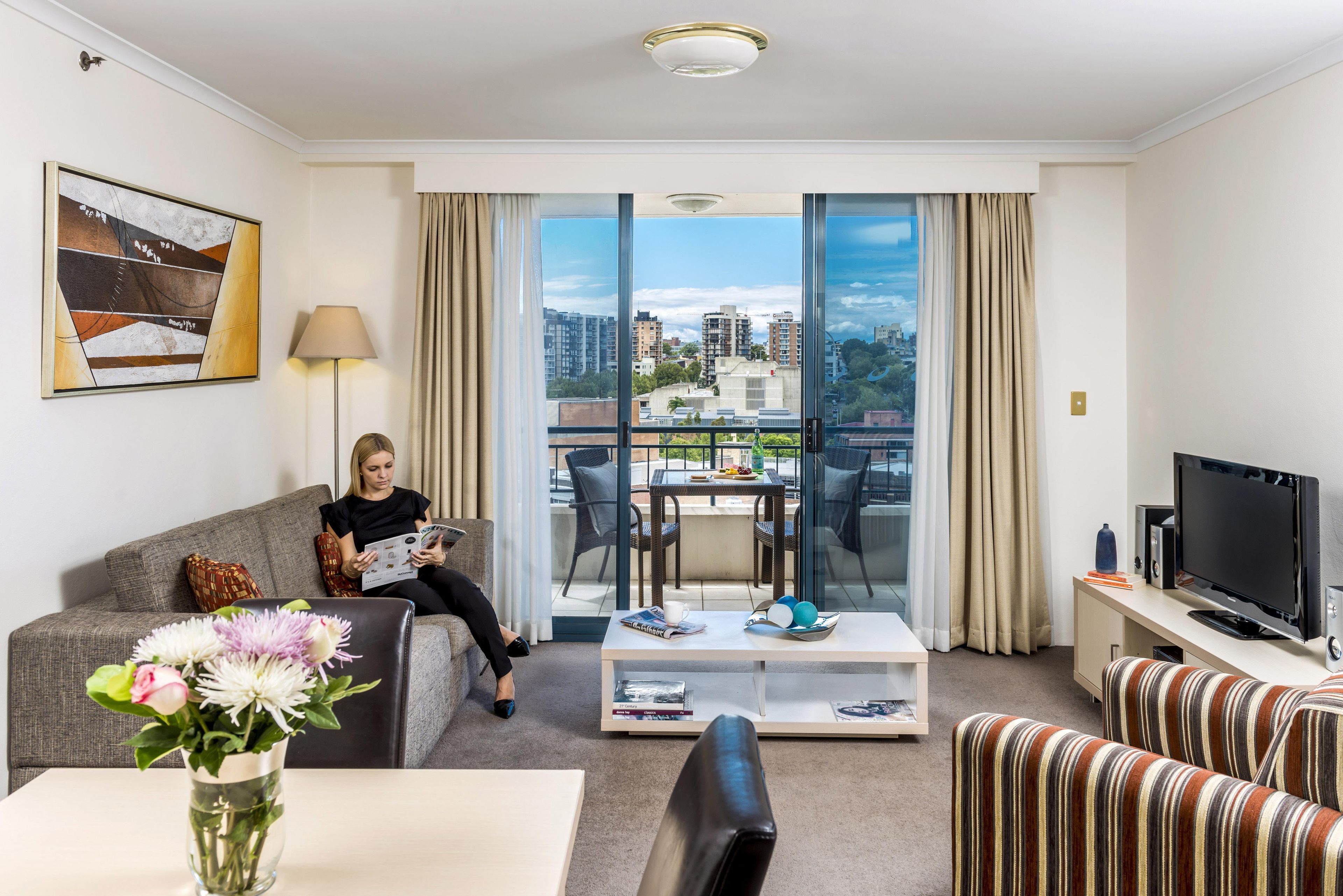 Una habitación de hotel moderna con un ventanal grande, un televisor de pantalla plana, un sofá, una mesa de centro y un comedor con una mesa y sillas.