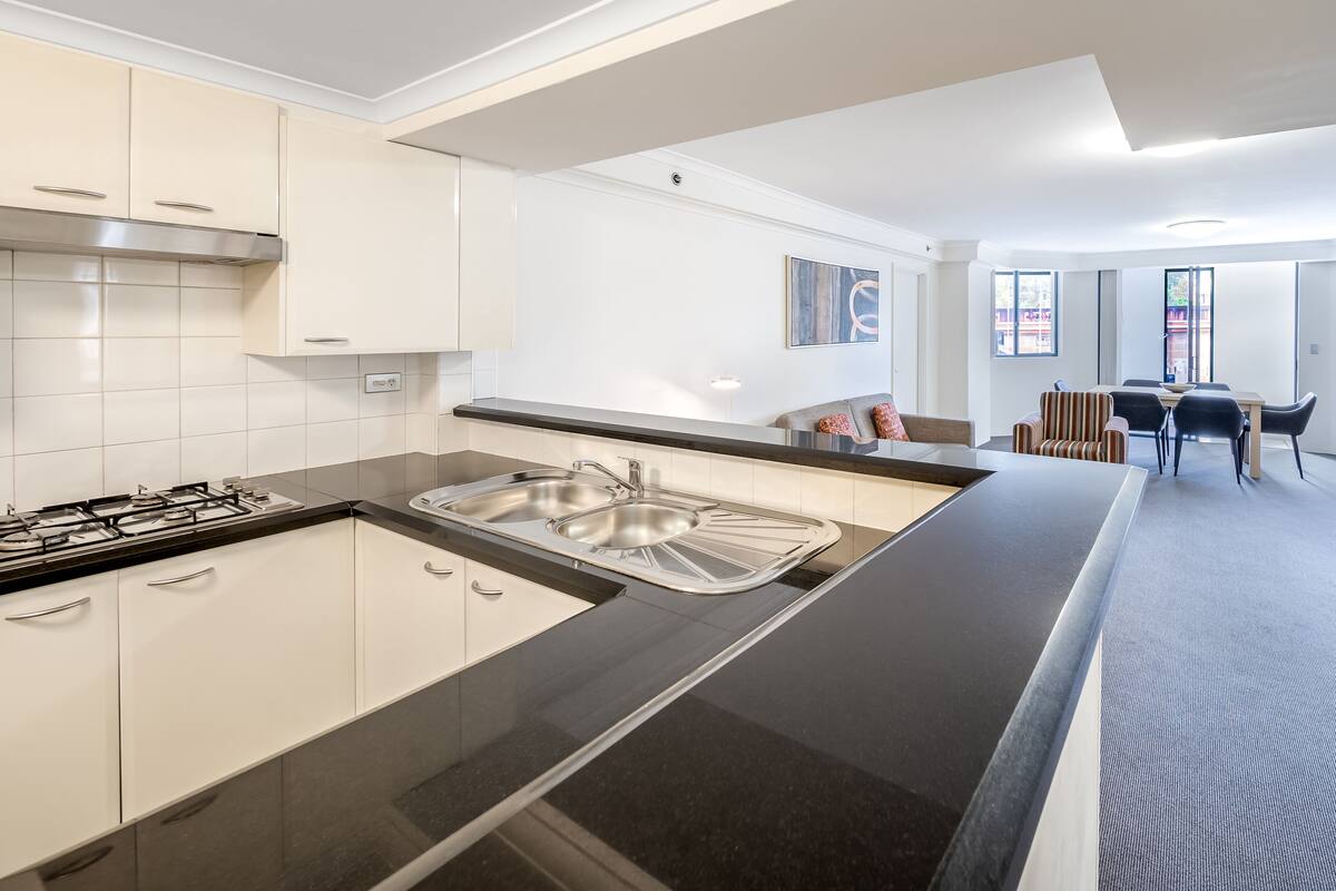 A modern kitchen with a gas stove, stainless steel sink, and white cabinetry. There is a dining area with a table and chairs in the background.
