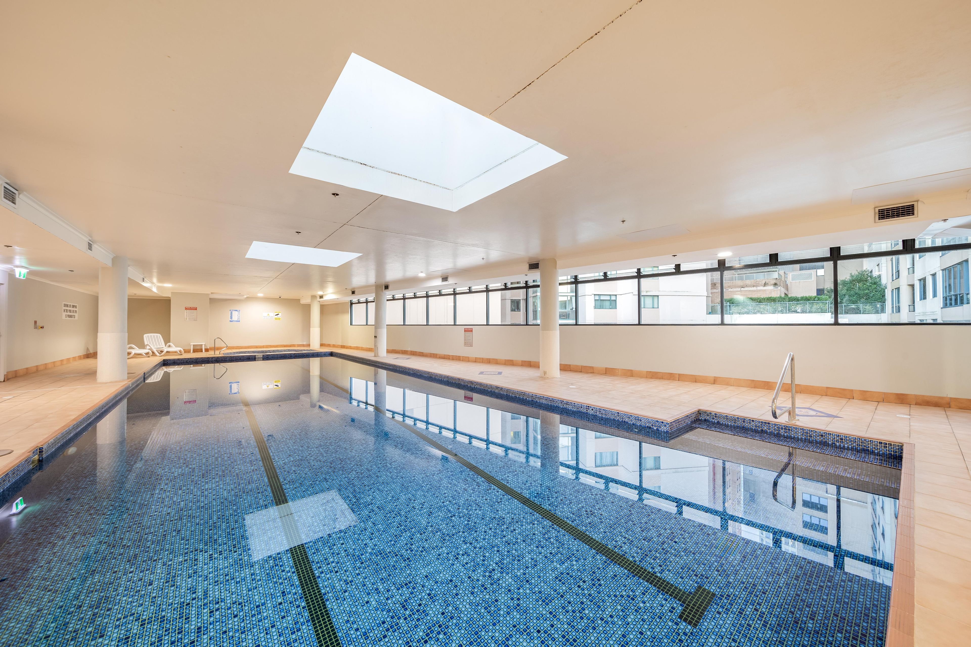 A large indoor swimming pool with clear blue tiles, a tiled deck area, and a view of buildings outside through large windows.