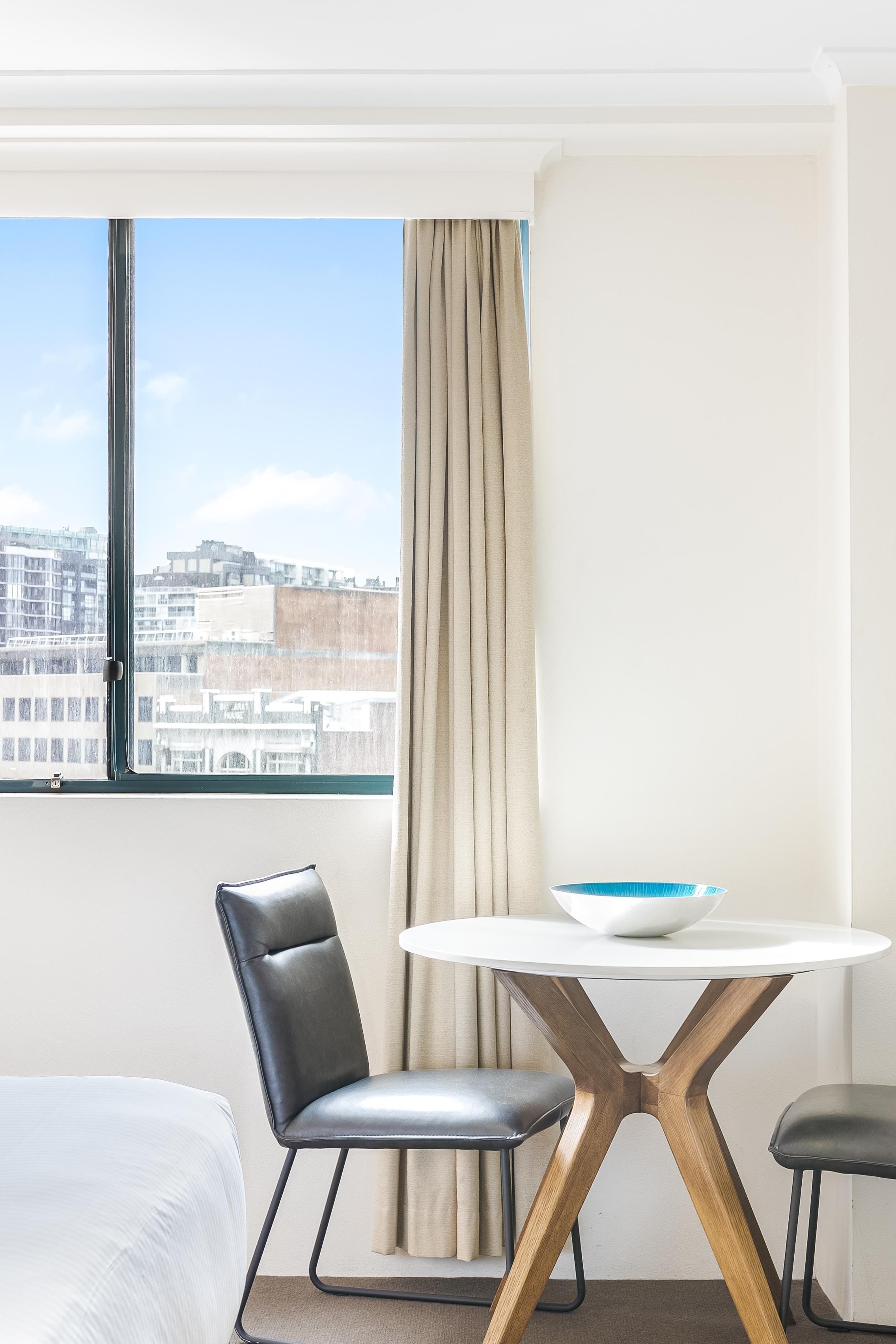 A hotel room with a bed, a chair, a table with a bowl, a window with a view of buildings, and a curtain.