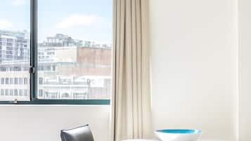A hotel room with a bed, a chair, a table with a bowl, a window with a view of buildings, and a curtain.