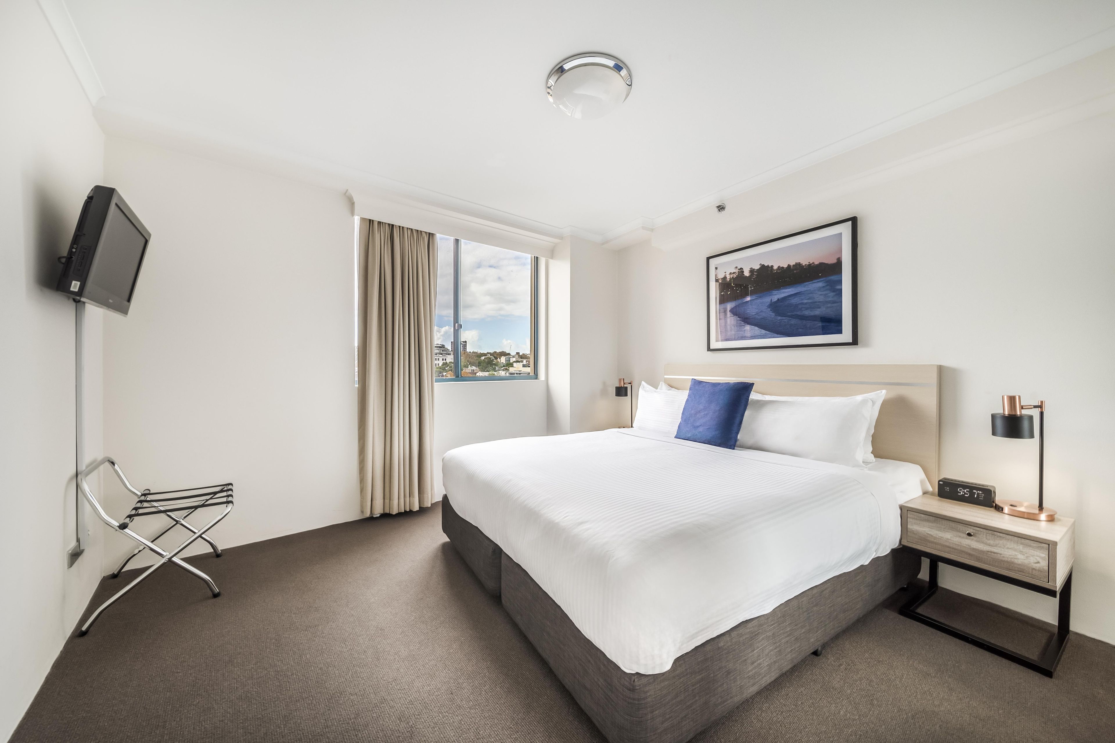 Habitación de hotel con una cama grande, televisión montada en la pared, ventana con cortinas, mesita de noche con lámpara y un cuadro enmarcado en la pared.