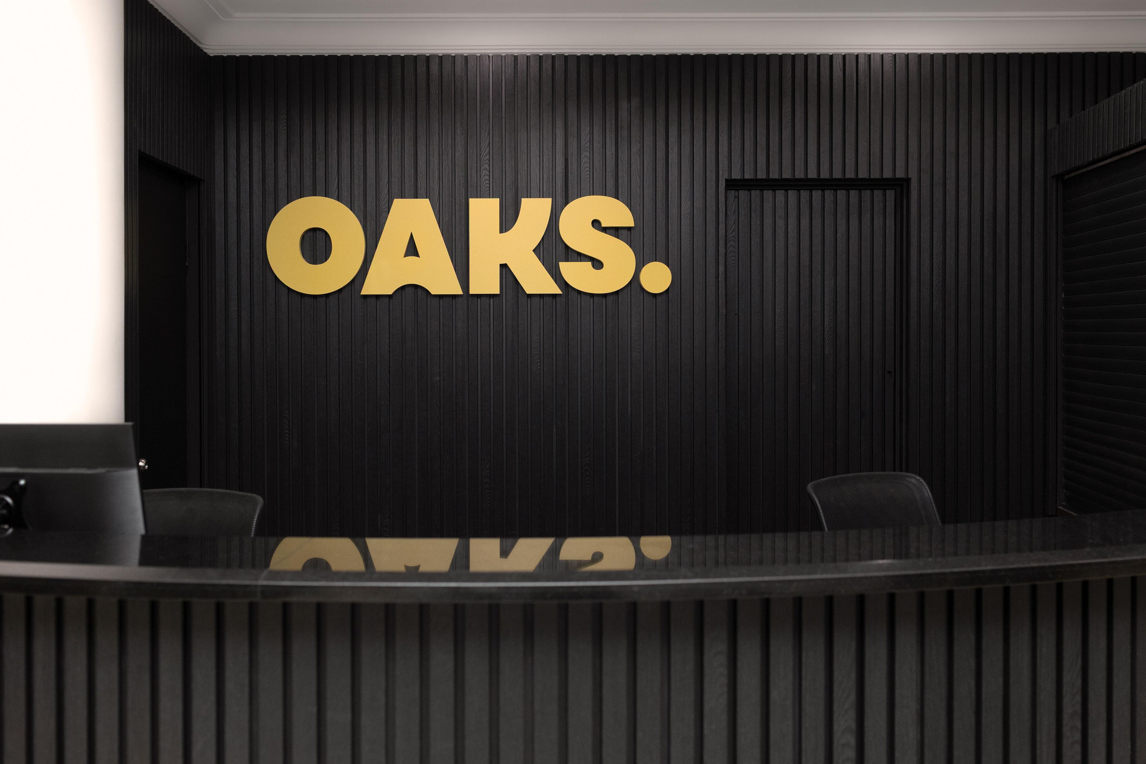 A reception desk with a dark wood panel wall and large gold letters spelling 'OAKS.'