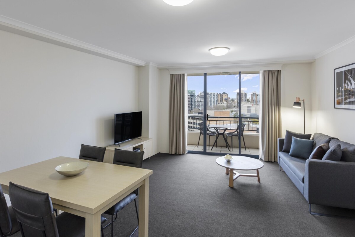 Habitación de hotel con mesa de comedor, sofá, televisión y balcón con vista a la ciudad.