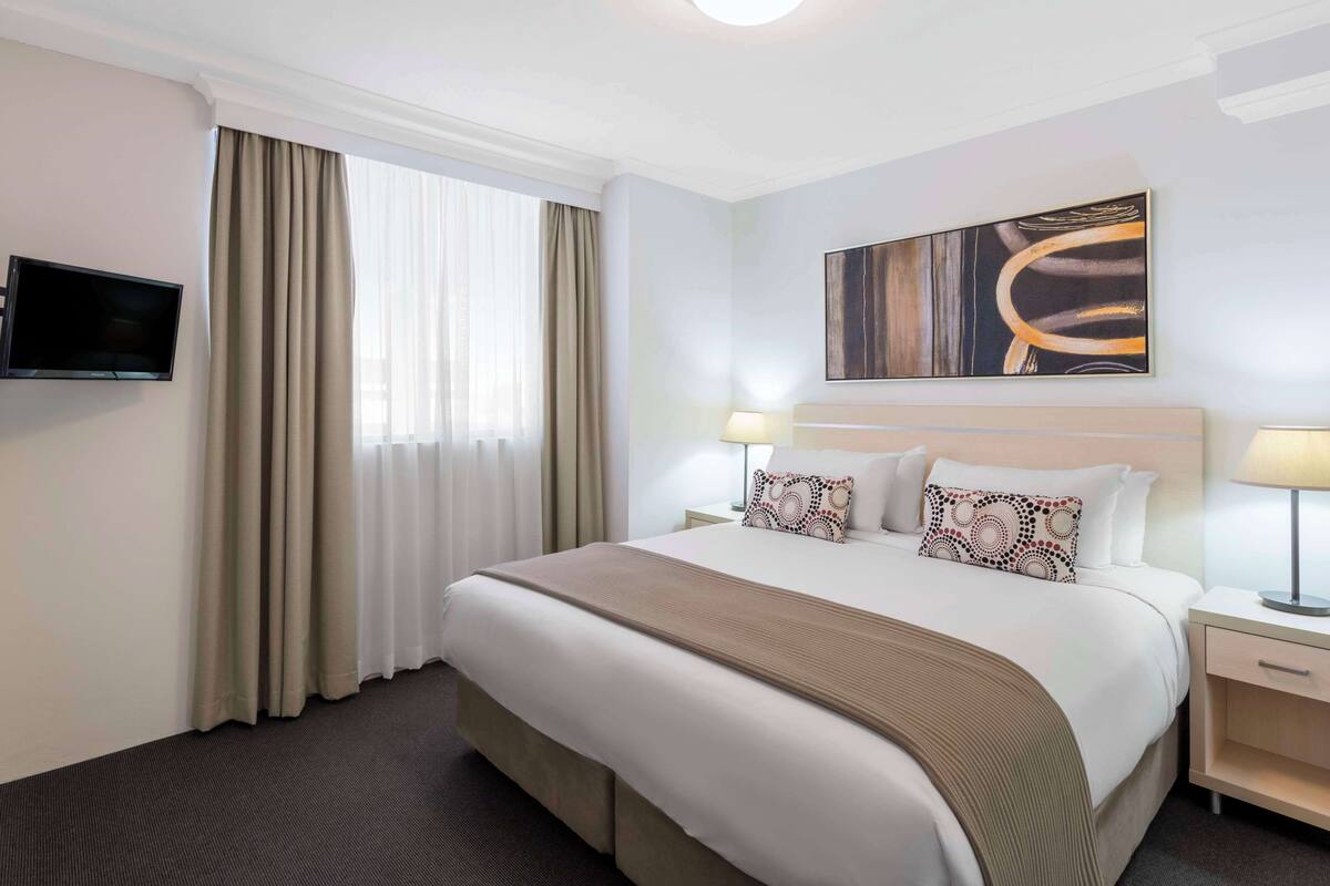 A hotel room with a large bed, two bedside tables with lamps, a flat-screen TV mounted on the wall, and a window with curtains.
