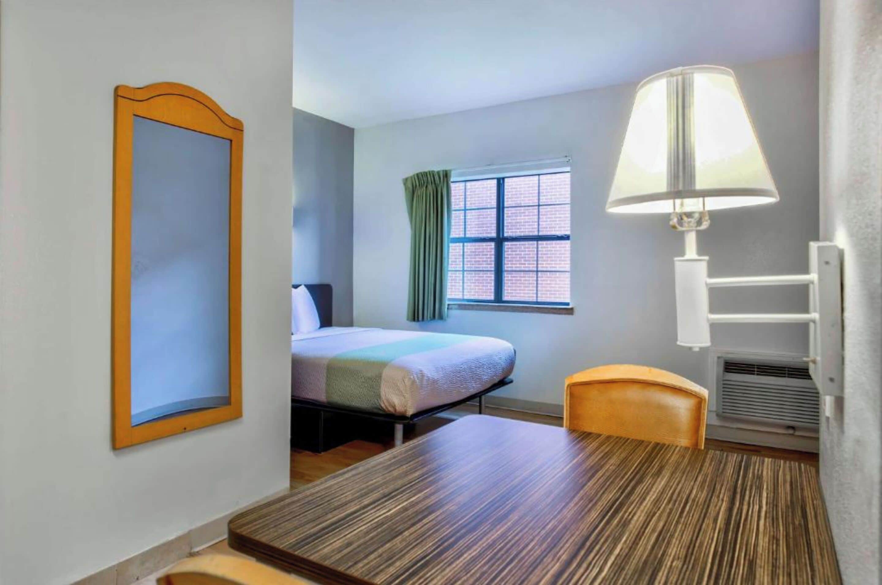 A hotel room with a bed, a desk, a chair, a window with curtains, a wall-mounted lamp, and a radiator.