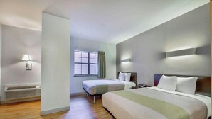 Ein Hotelzimmer mit einem großen Bett, einem kleineren Bett, einem Fenster mit Vorhängen, einer Wandlampe und einem Heizkörper.
