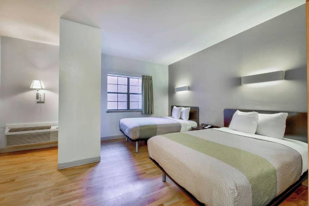 Una habitación de hotel con una cama grande, una cama más pequeña, una ventana con cortinas, una lámpara de pared y un calefactor.
