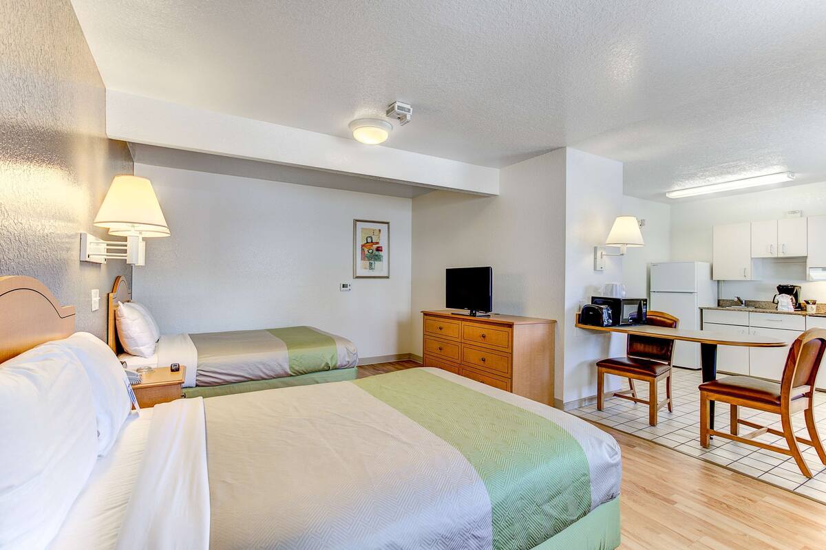 A hotel room with two beds, a desk, a chair, a television, and a kitchenette.