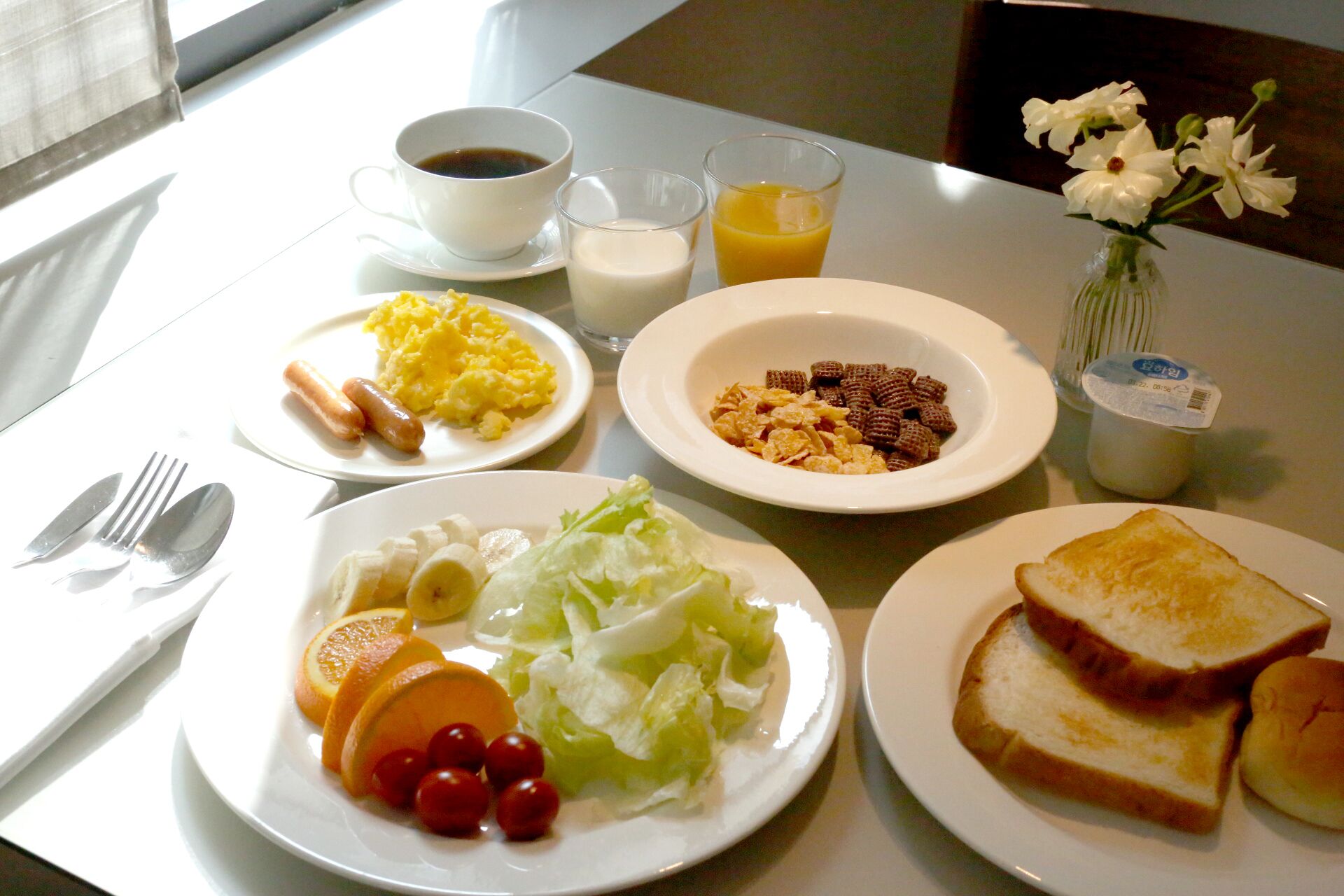 A breakfast table with plates of eggs, sausages, toast, and fruit, along with cups of coffee and orange juice.