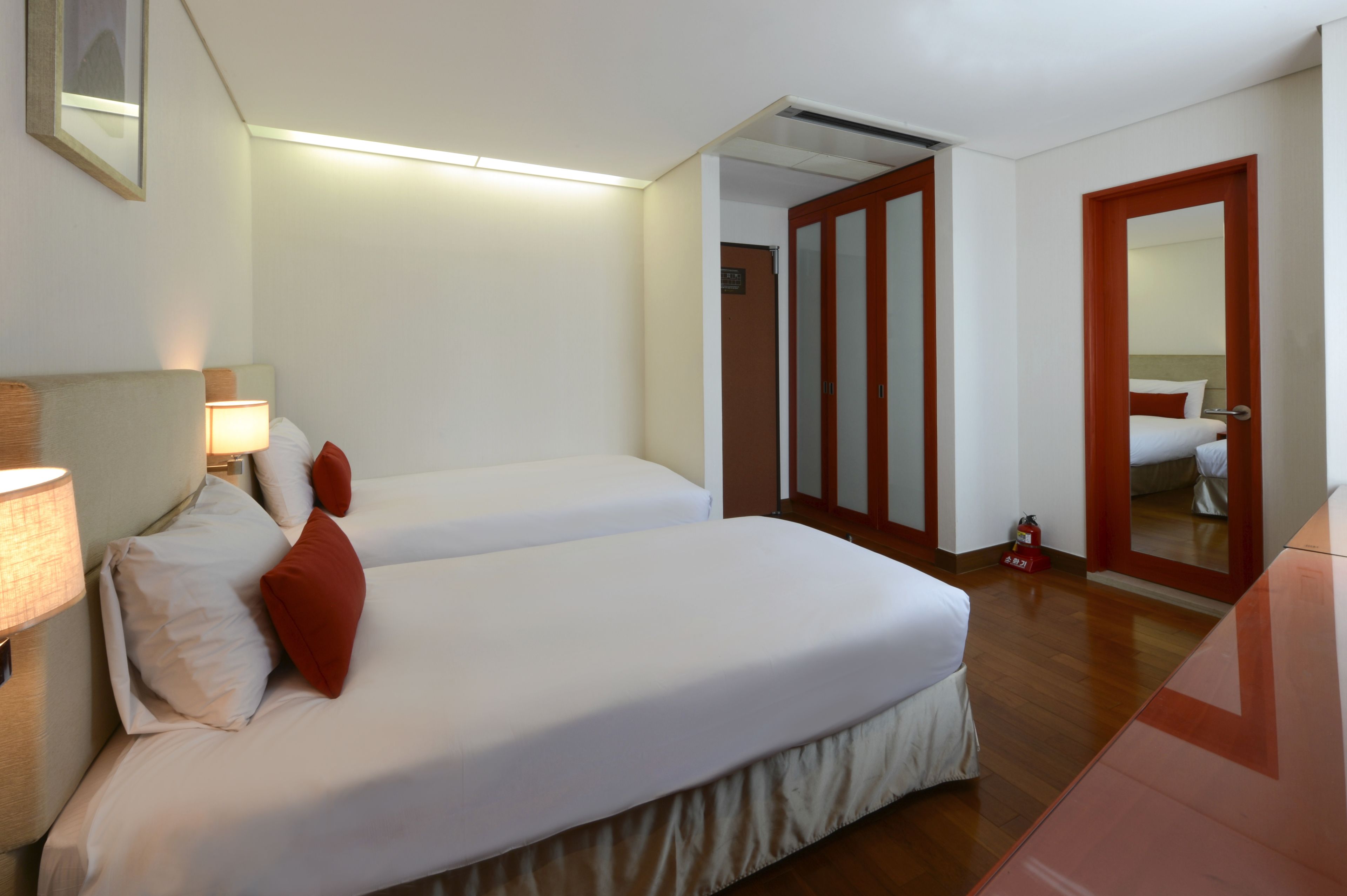 A hotel room with two beds, a bedside lamp, a fire extinguisher, and a mirror on the door.