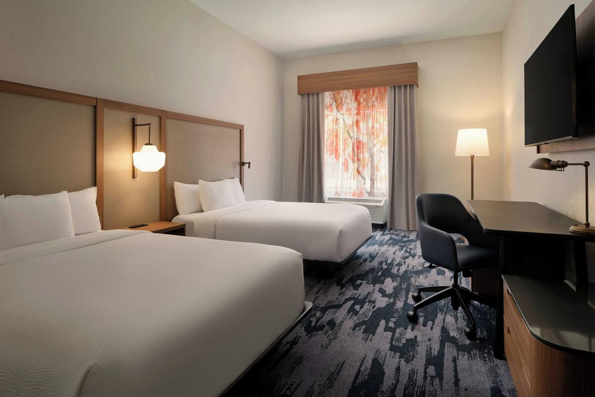 A hotel room with two beds, a desk, a chair, a TV, and a window with curtains.