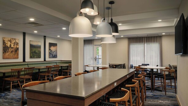 A hotel lobby with a long communal table, multiple chairs, hanging pendant lights, and artwork on the walls.