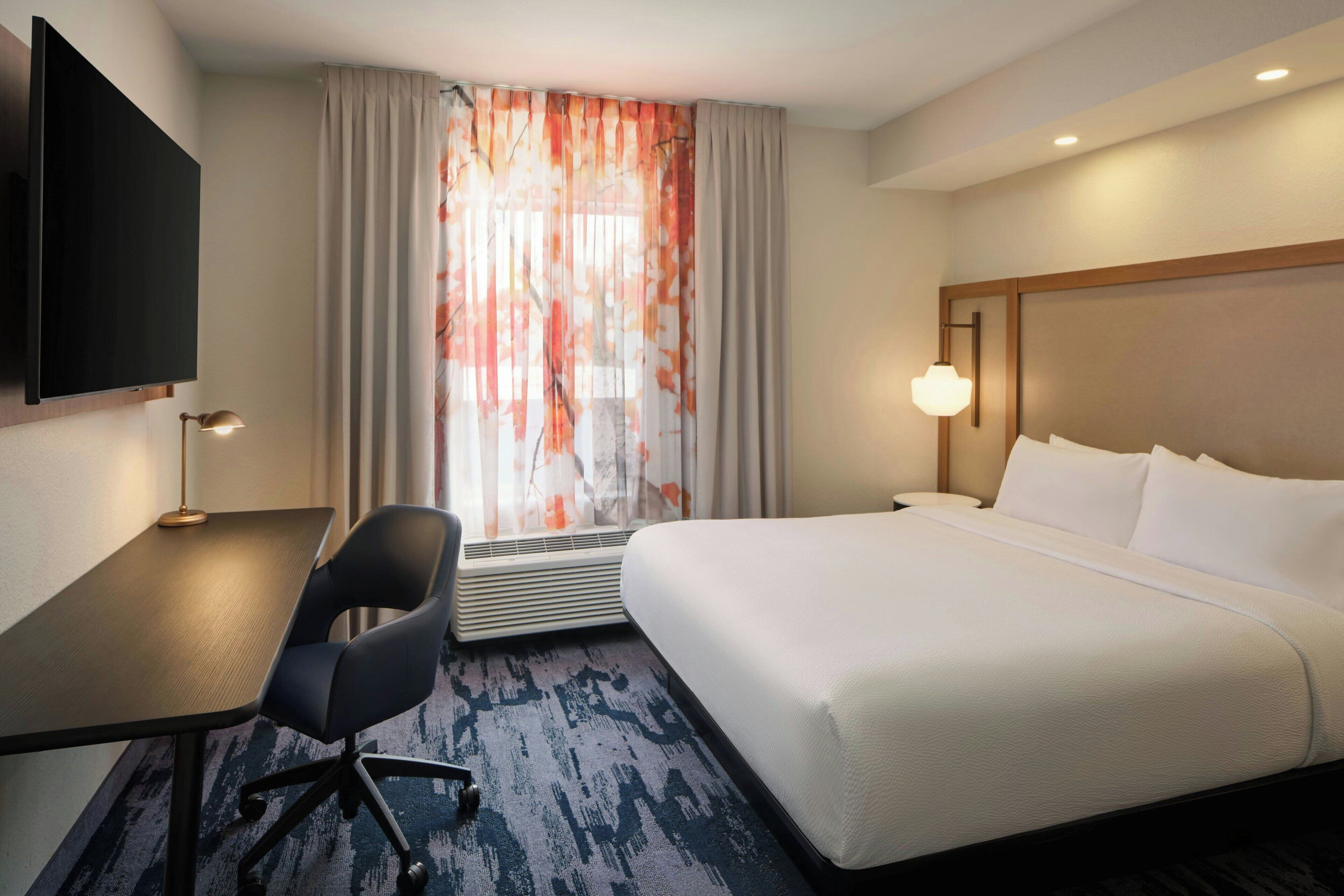A hotel room with a bed, a desk, a chair, a TV, and a window with curtains.