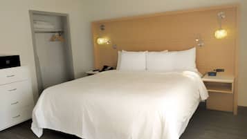 A hotel room with a large bed, a bedside table with a lamp, a nightstand with a clock, a wardrobe, and a desk with a computer.