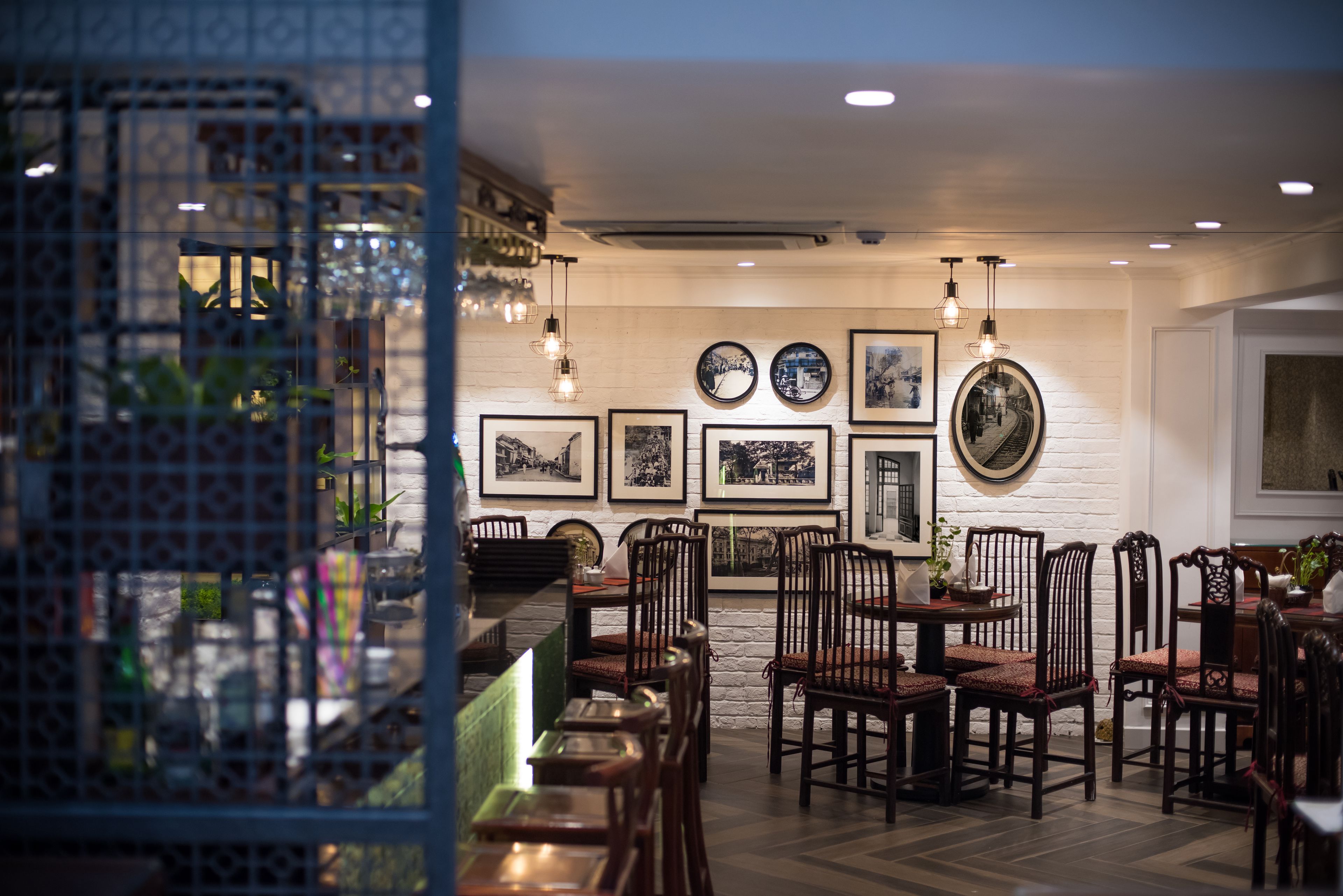 Un restaurant avec des chaises et des tables en bois, un mur en briques blanches orné de tableaux encadrés et des lampes suspendues.