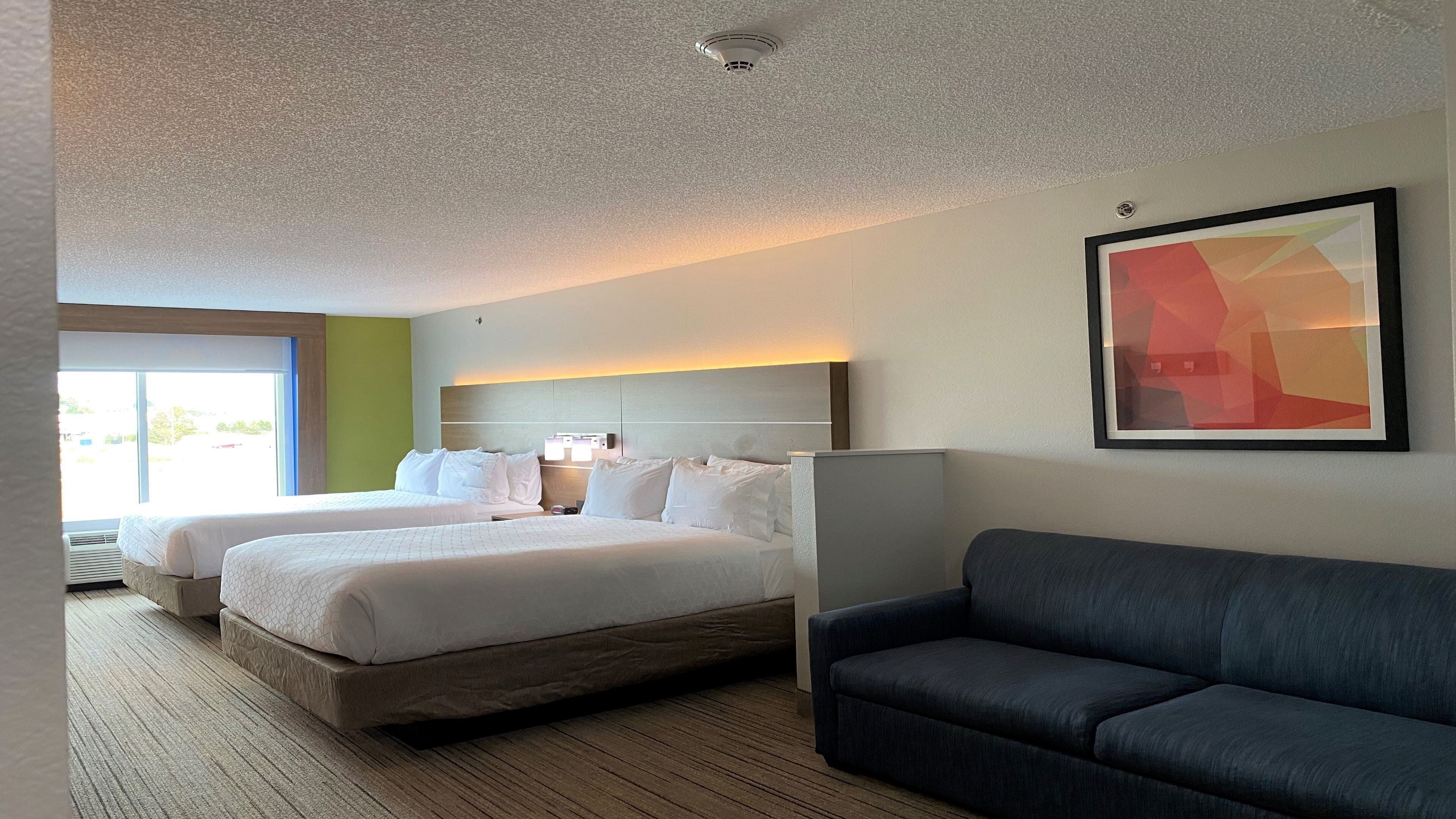 Una habitación de hotel con dos camas, un sofá gris, un ventanal grande y un cuadro abstracto enmarcado.