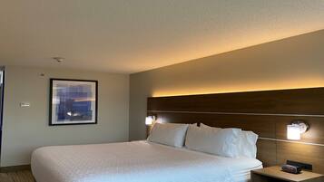 A hotel room with a large bed, a headboard with built-in lighting, a framed picture on the wall, and a bedside table with a clock.
