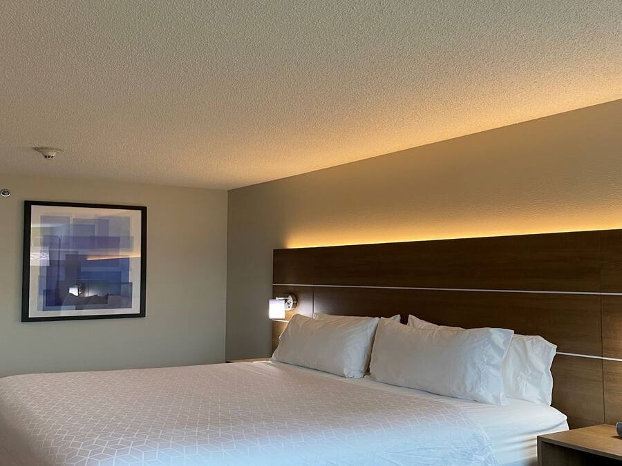 A hotel room with a large bed, a headboard with built-in lighting, a framed picture on the wall, and a bedside table with a clock.