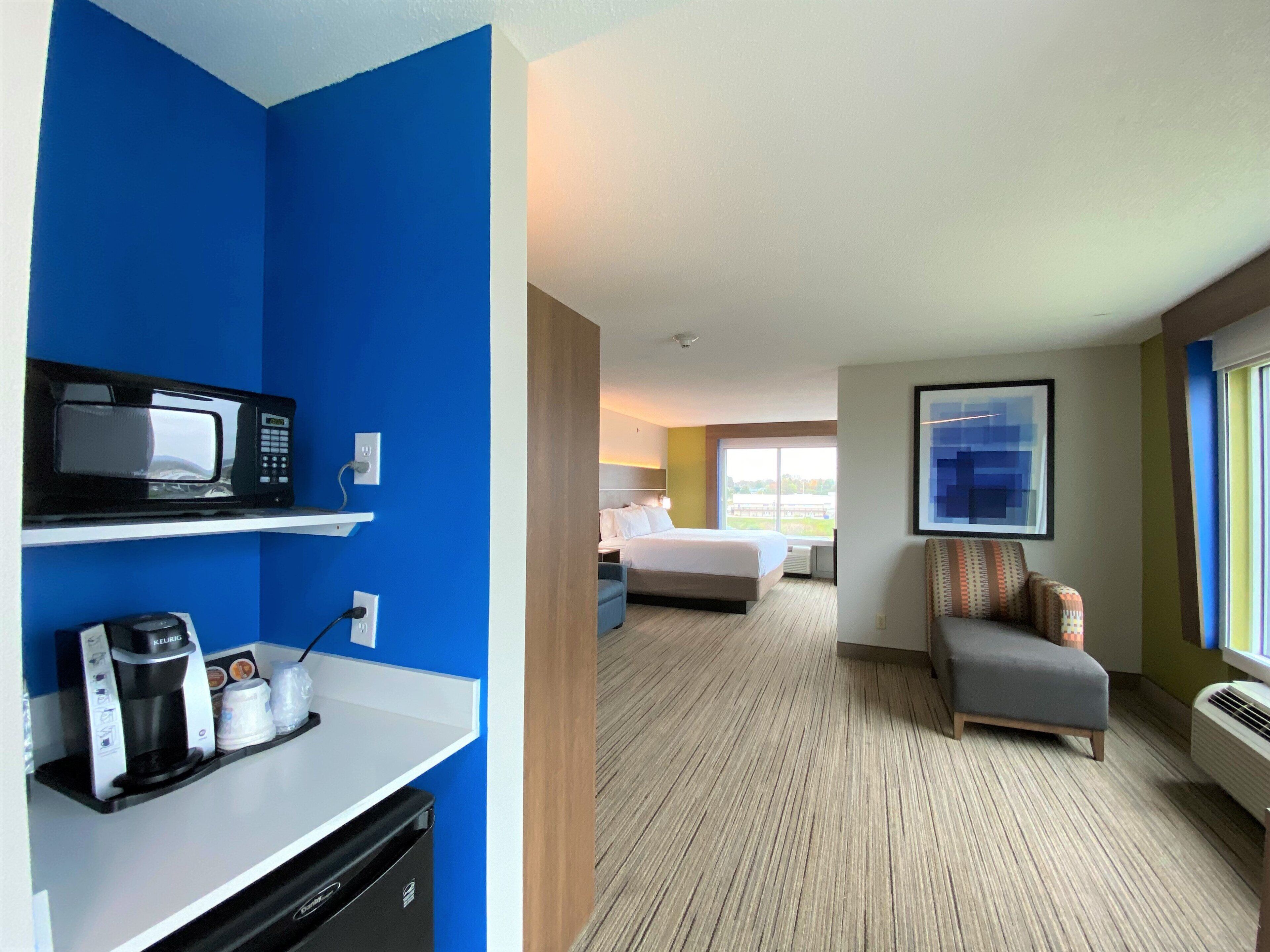 A hotel room with a kitchenette, a bed, a seating area, and a large window.