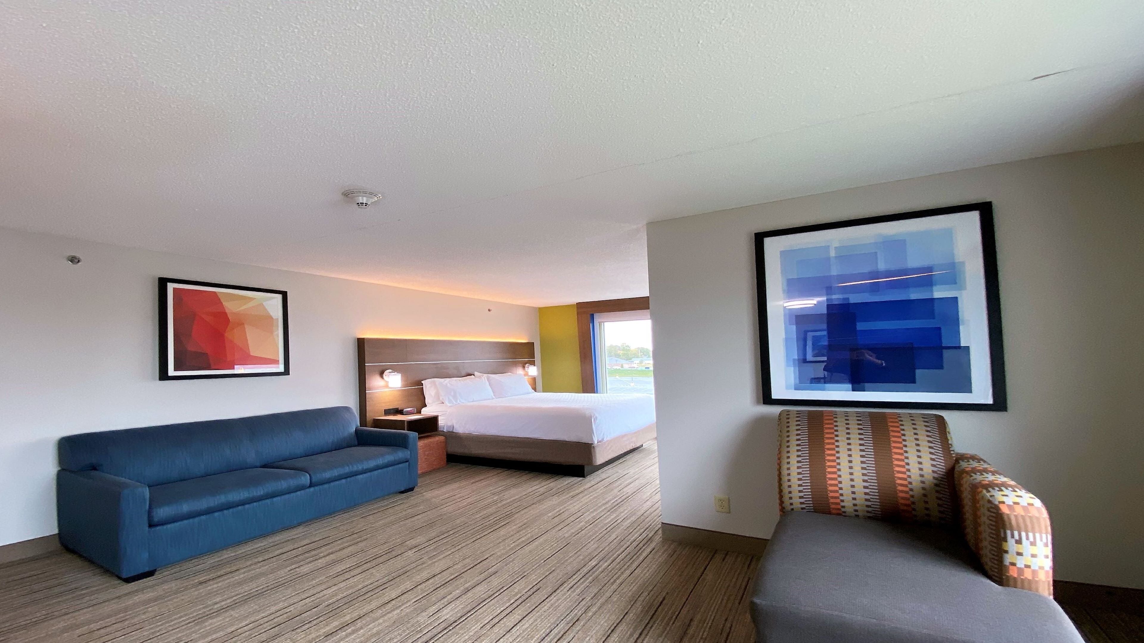 Una habitación de hotel con una cama, un sofá azul, un sillón estampado y dos cuadros abstractos.