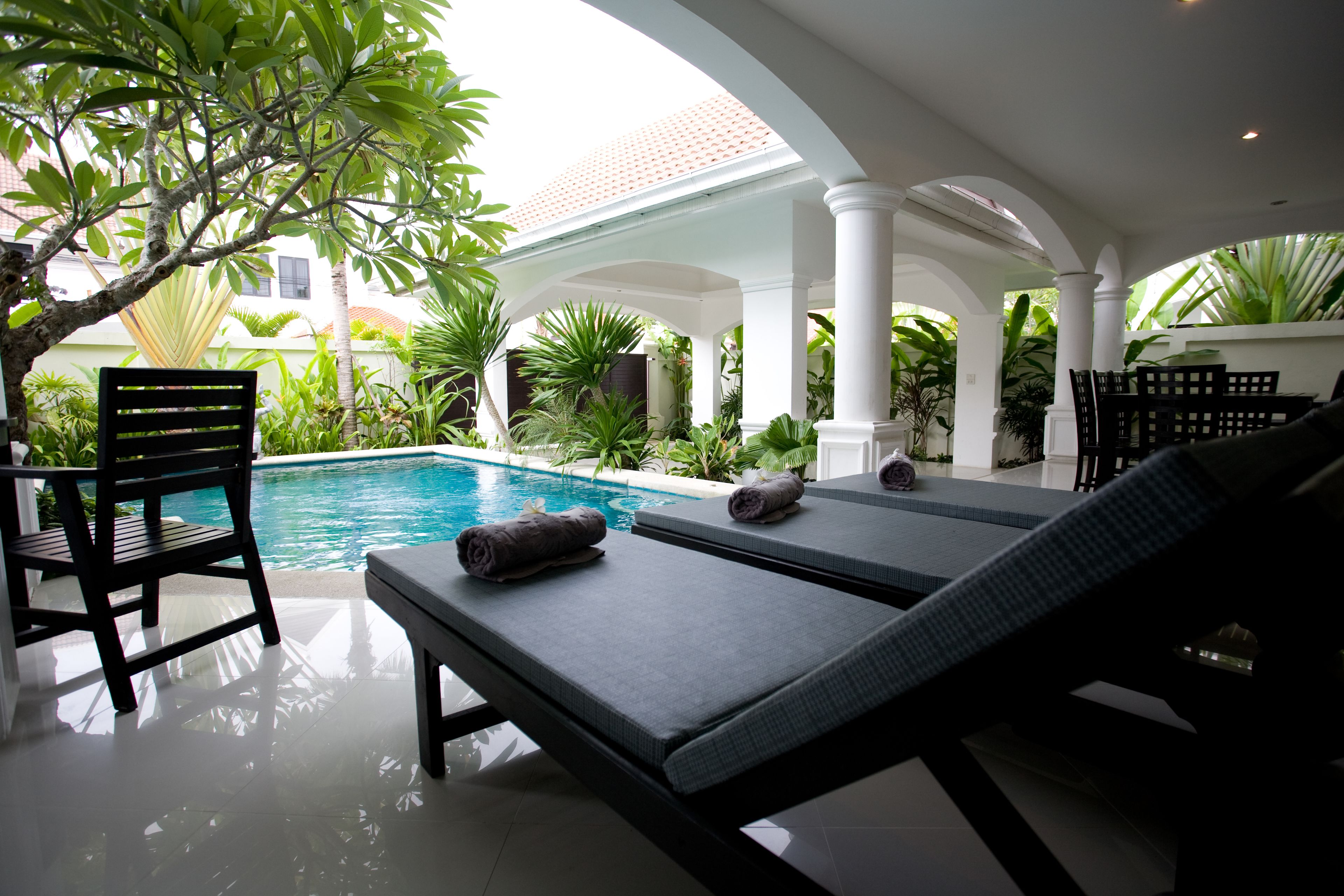 A poolside area with lounge chairs and a chair, surrounded by greenery and a tiled roof.