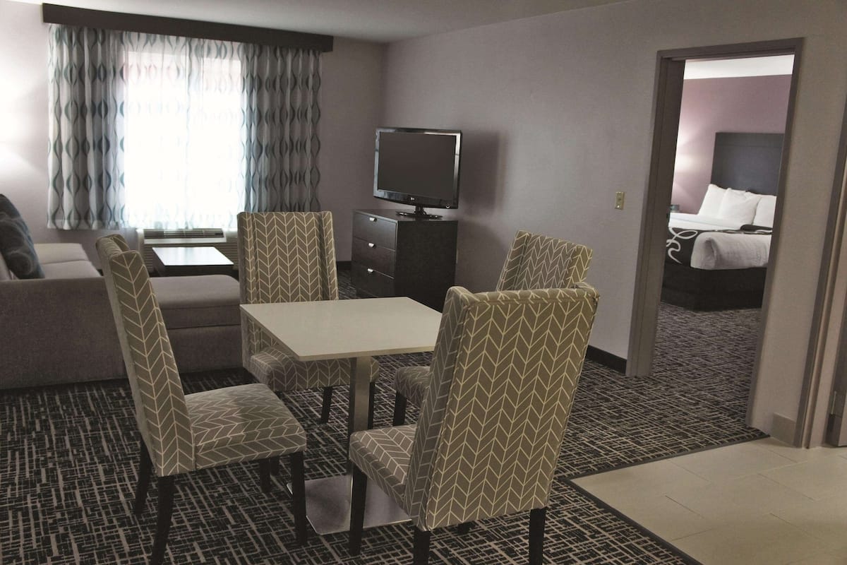 Habitación de hotel con comedor, televisor, cama y baño visible a través de una puerta abierta.