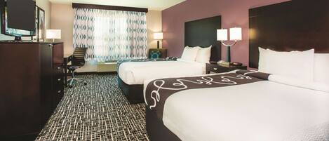 A hotel room with two beds, a desk, a television, and a window with patterned curtains.