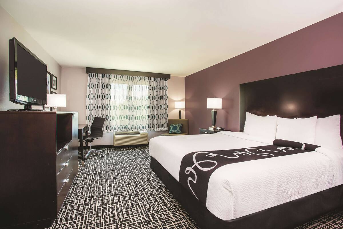Habitación de hotel con una cama grande, un escritorio con televisor, una silla y una alfombra estampada.