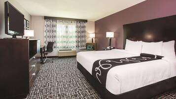 A hotel room with a large bed, a desk with a television, a chair, and a patterned carpet.