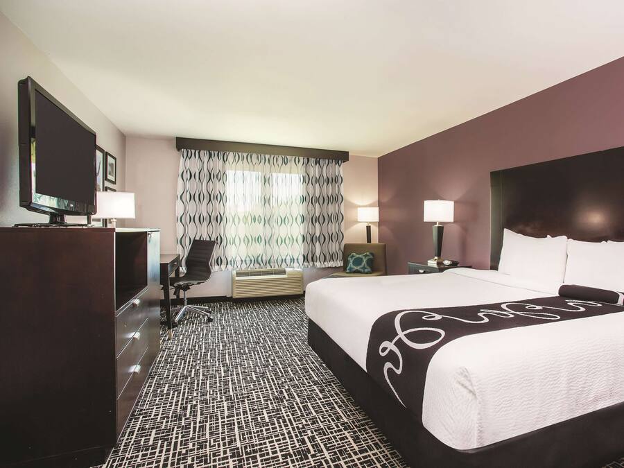 A hotel room with a large bed, a desk with a television, a chair, and a patterned carpet.