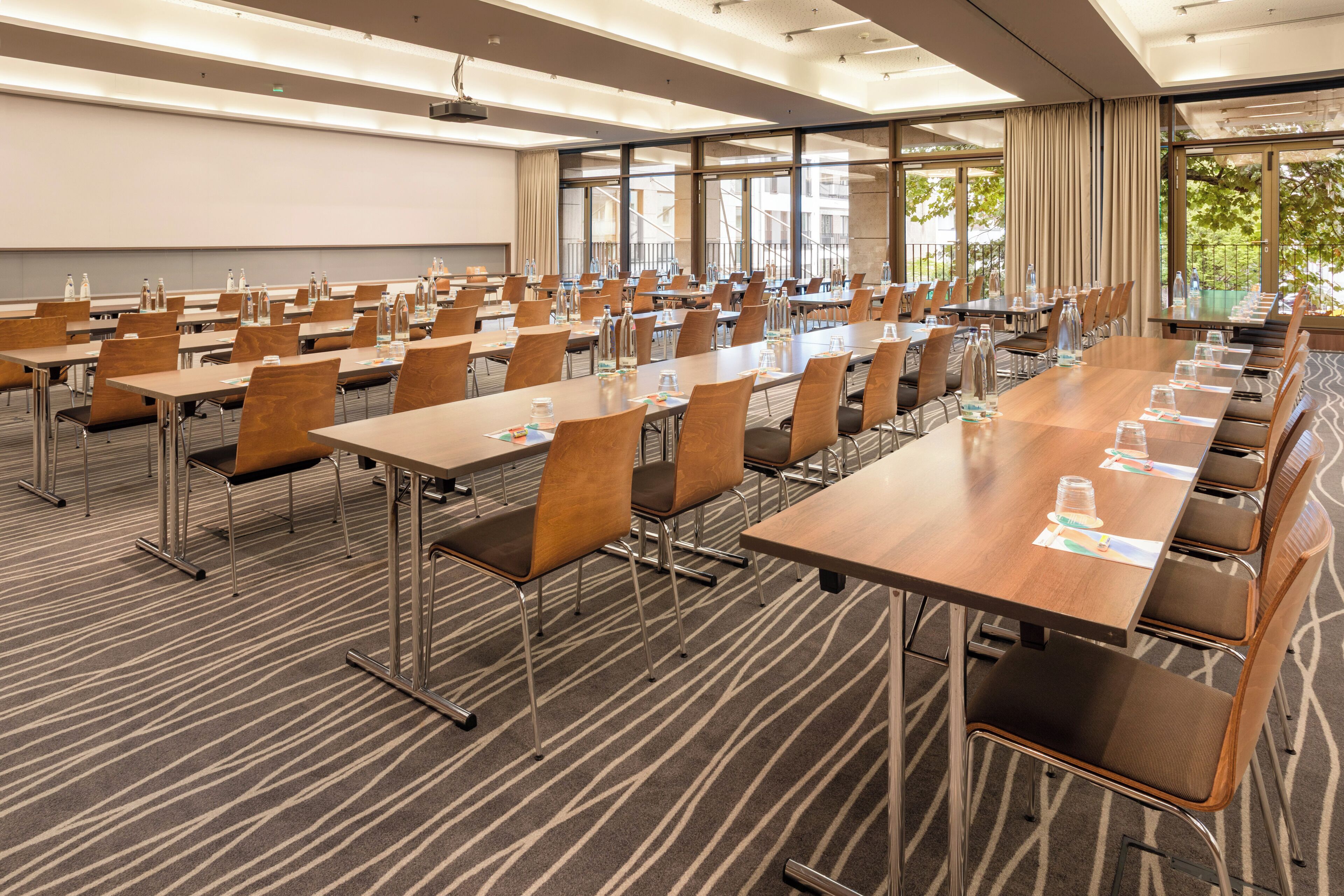 A conference room with multiple tables and chairs, a projector screen, and large windows with curtains.