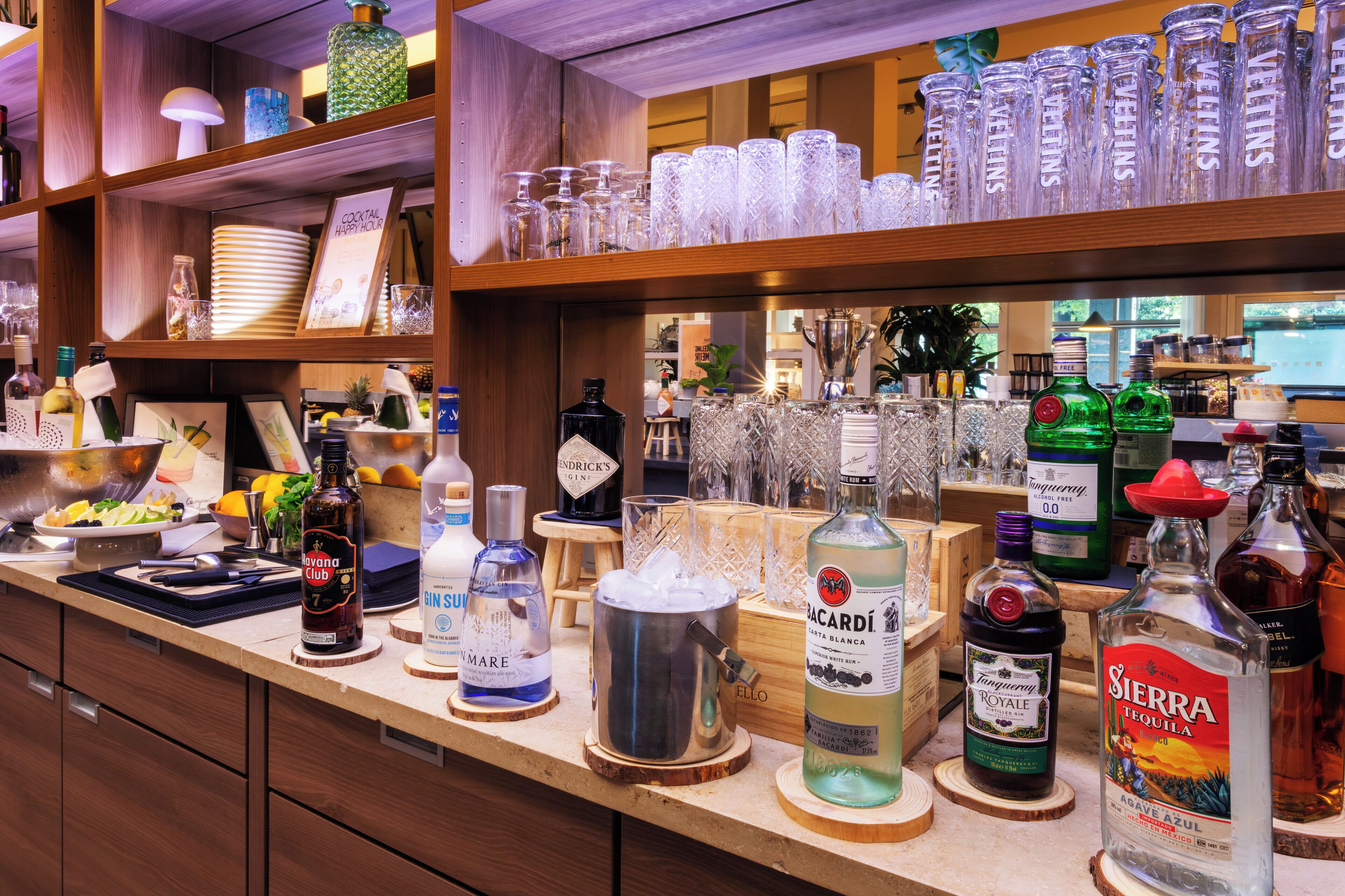 Zona de bar con diversas botellas de alcohol, vasos y un cartel que anuncia una hora feliz de cócteles.
