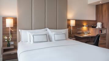 Una habitación de hotel moderna con una cama grande, mesitas de noche con lámparas, un escritorio con cafetera y un cabecero de madera.