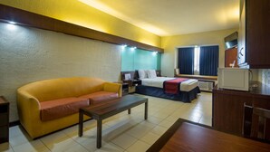 A hotel room with a yellow sofa, a wooden coffee table, a bed with a blue blanket, a nightstand, a desk, a microwave, and a window with curtains. - Microtel by Wyndham Eagle Ridge (General Trias)