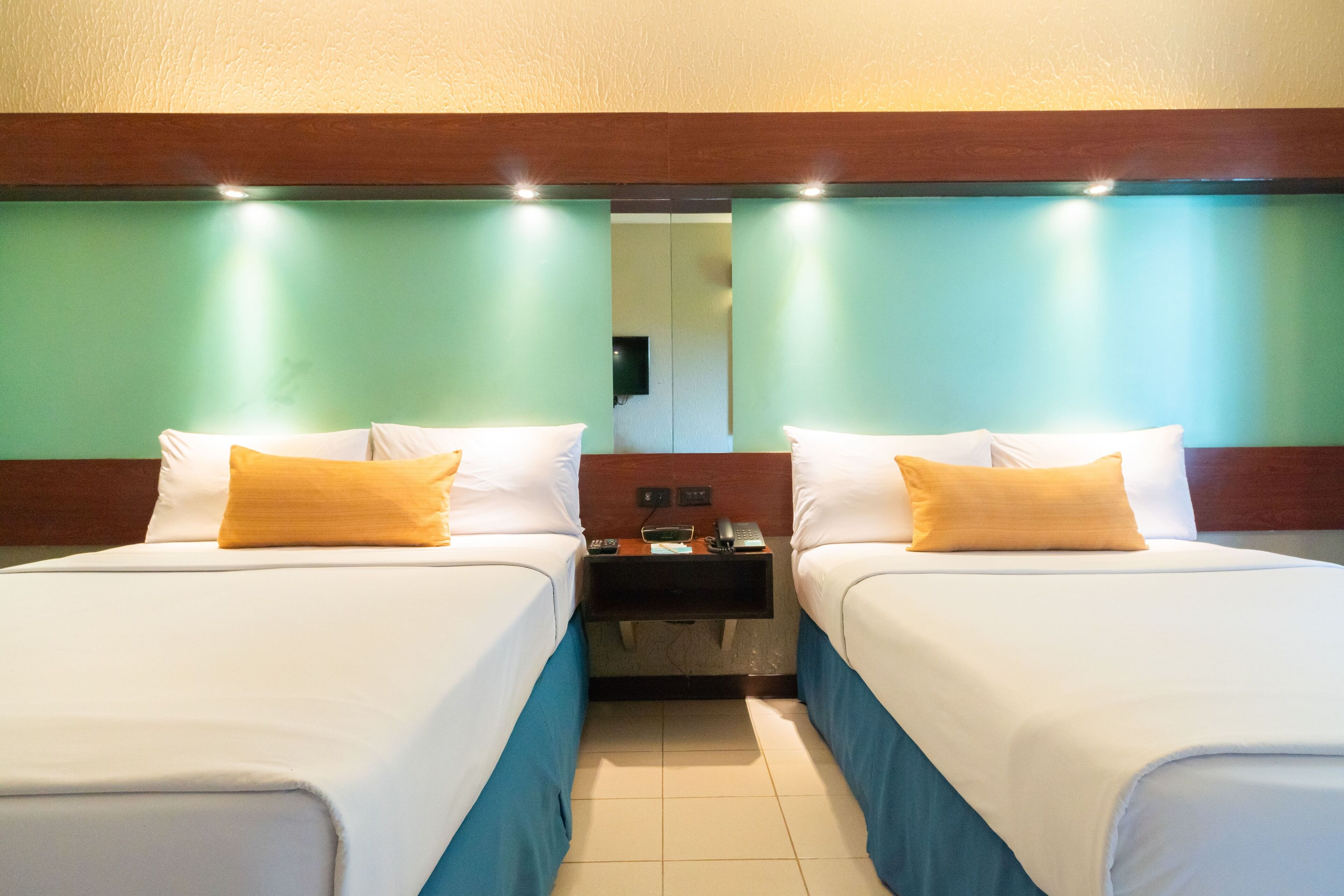Habitación de hotel con dos camas, cada una con ropa blanca y una almohada amarilla. Entre las camas hay una mesita de noche de madera con un teléfono y una lámpara pequeña.