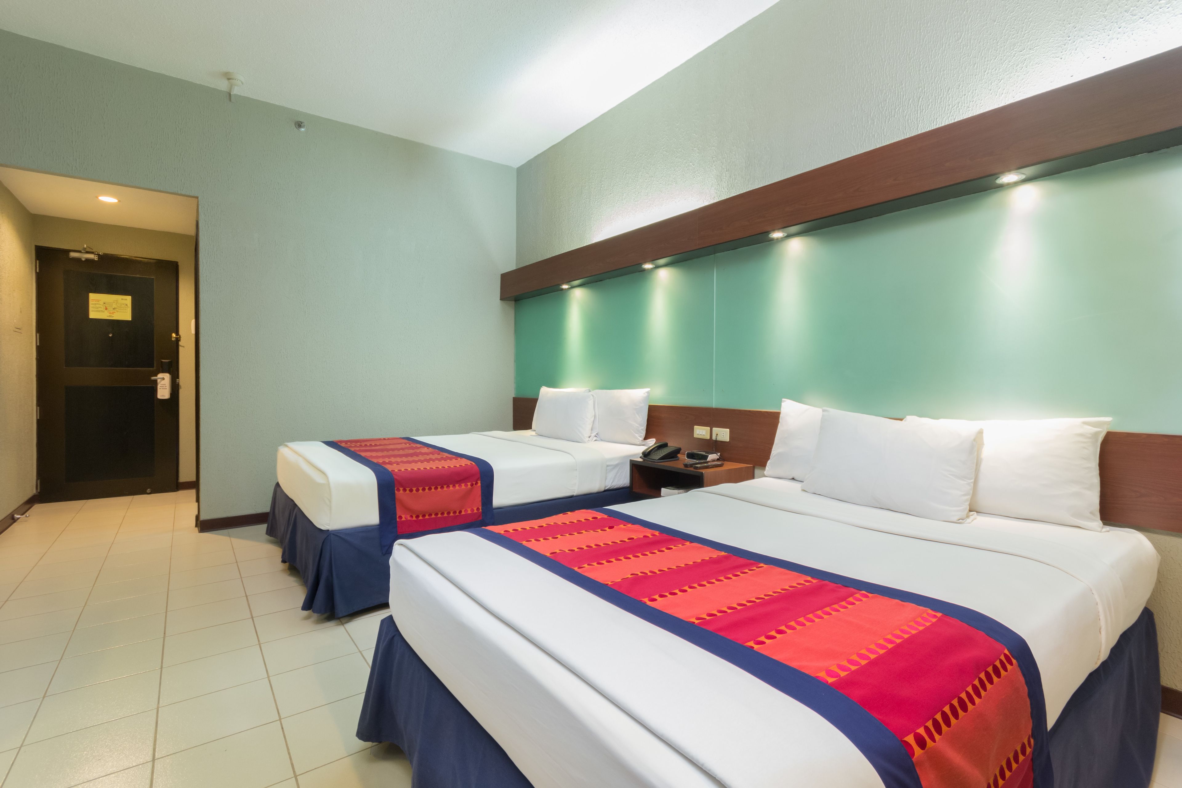 A hotel room with two beds, a wooden headboard, a nightstand, and a door leading to another room.